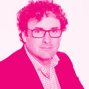 Malcom Fraser has curly hair, large rectangular glasses and wears a check shirt and blazer while staring into the camera. Photo uses a hot pink filter.